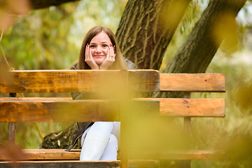 Image showing A young beautiful girl sits on a bench in an autumn park and looks cheerfully into the frame