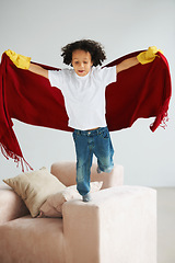 Image showing Fly, children and a boy superhero in a costume on a sofa in the living room of his home for playing a game. Jumping, growth and fantasy with a young kid in a cape for justice or safety cosplay