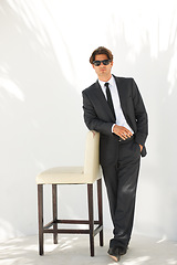 Image showing Portrait, fashion and a business man with a chair in studio on a white background for corporate style. Handsome, sunglasses and formal with a confident young professional employee leaning on a seat