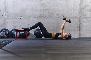 Image showing A fit woman is lying on the gym floor, performing arm exercises with dumbbells and showcasing her dedication and strength.