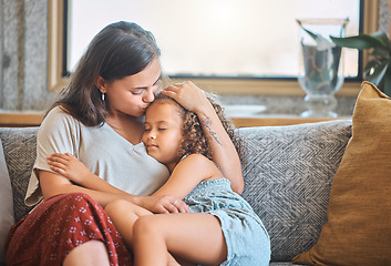 Image showing Loving mother kissing her daughter on the forehead as she falls asleep in her arms while sitting on the couch at home. Tired little girl falling asleep feeling safe and loved in mothers arms