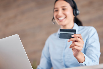 Image showing Happy young mixed race call center agent holding a credit card in her hand and working on a laptop while wearing a headset and answering calls in an office at work