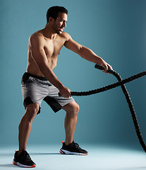 Image showing Handsome young hispanic man training with battle ropes in studio isolated against a blue background. Mixed race shirtless male athlete exercising or working out to increase his strength and fitness