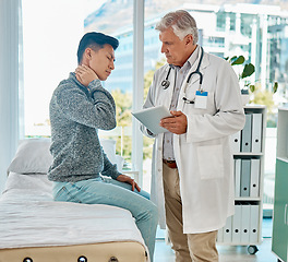 Image showing Mature medical doctor standing and using a digital tablet while checking on his injured asian patient in a clinic. Young mixed race man suffering from a painful neck in a hospital. Tension and sprain