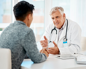 Image showing Mature caucasian medical doctor sitting with his patient during a consultation in a clinic and talking. Healthcare professional using hand gestures while discussing treatment with sick mixed race man