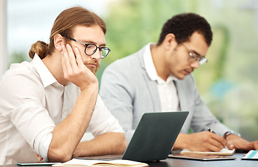 Image showing When will this day end. a young businessman looking bored while working on a laptop in an office with his colleague in the background.