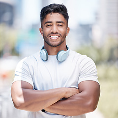 Image showing Fitness man wearing headphones around his neck while looking at the camera and smiling. Cheerful male runner standing with his arms crossed while out in the city for a workout