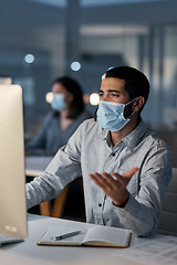 Image showing Communication can get you through the darkest days. a masked young man using a headset and computer late at night in a modern office.