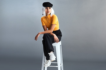 Image showing Do you know the history behind the baker boy hat. Studio shot of a beautiful young woman sitting on a stool against a grey background.