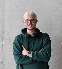 Image showing A man with blue hair, eyeglasses, and a green sweatshirt confidently poses with his arms crossed against a gray background, showcasing his fashionable and unique style.
