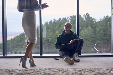 Image showing A young blond man in a modern office is sitting by the window, engrossed in his work on a laptop while talking to a female colleague