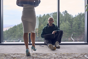 Image showing A young blond man in a modern office is sitting by the window, engrossed in his work on a laptop while talking to a female colleague