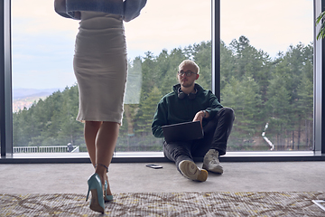 Image showing A young blond man in a modern office is sitting by the window, engrossed in his work on a laptop while talking to a female colleague