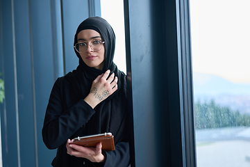 Image showing A hijab businesswoman stands by the window in a modern office holding a tablet in her hand showcasing her professionalism, technological prowess and entrepreneurial spiri