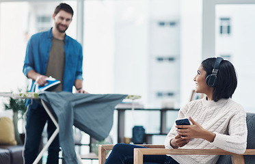 Image showing Teamwork makes the home work. a young woman using a smartphone and headphones while her husband irons clothing in the background.