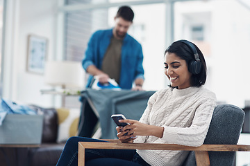 Image showing Today it’s hubby’s turn, tomorrow it’s mine. a young woman using a smartphone and headphones while her husband irons clothing in the background.