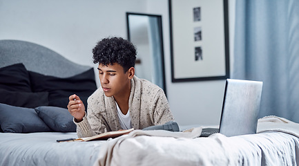 Image showing 2020 had a slight change in plans. a young man writing in a notebook and using a laptop on his bed at home.