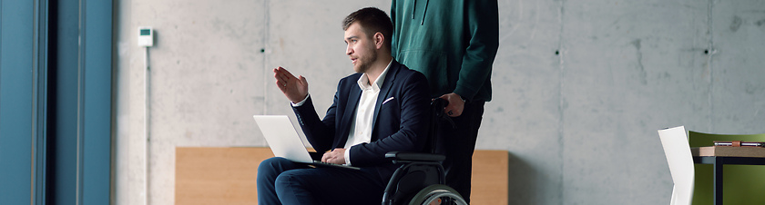 Image showing Wide crop of businessman in a wheelchair in a fashionable office using a laptop while behind him is his business colleague who gives him support