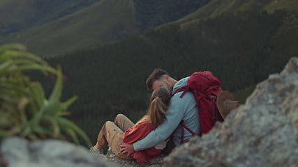 Image showing Mountain, sunset or couple kiss to relax outdoors together on adventure in nature from back for view. Love, hiking or romantic man hugging woman sitting on cliff on camping holiday, vacation or break