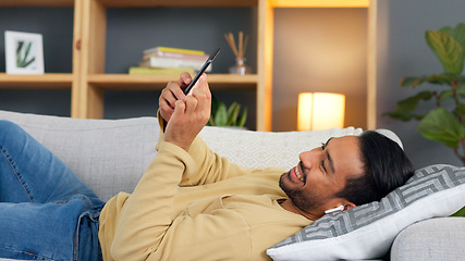 Image showing Man, phone and earphones to relax on sofa, texting or reading with smile, contact or music in home living room. Guy, chat and smartphone for audio streaming subscription, video or comic meme on couch