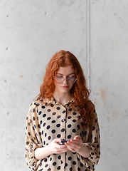 Image showing A young entrepreneur with captivating orange hair is striking a confident pose while holding a smartphone in front of a stylish gray wall.