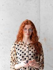 Image showing A young entrepreneur with captivating orange hair is striking a confident pose while holding a smartphone in front of a stylish gray wall.