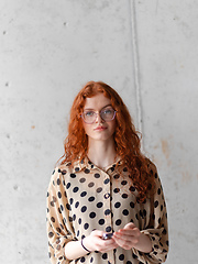 Image showing A young entrepreneur with captivating orange hair is striking a confident pose while holding a smartphone in front of a stylish gray wall.