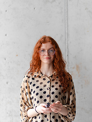 Image showing A young entrepreneur with captivating orange hair is striking a confident pose while holding a smartphone in front of a stylish gray wall.