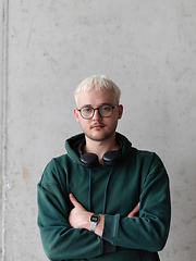 Image showing A man with blue hair, eyeglasses, and a green sweatshirt confidently poses with his arms crossed against a gray background, showcasing his fashionable and unique style.