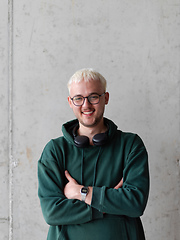 Image showing A man with blue hair, eyeglasses, and a green sweatshirt confidently poses with his arms crossed against a gray background, showcasing his fashionable and unique style.