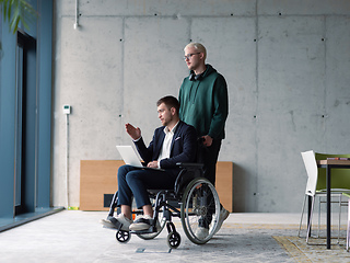 Image showing A businessman in a wheelchair in a fashionable office using a laptop while behind him is his business colleague who gives him support