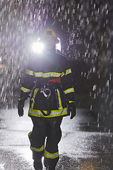 Image showing A determined female firefighter in a professional uniform striding through the dangerous, rainy night on a daring rescue mission, showcasing her unwavering bravery and commitment to saving lives.