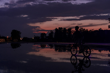 Image showing Lonely children silhouette on bike, boy riding bicycle on reflective water. Background beautiful sunset.