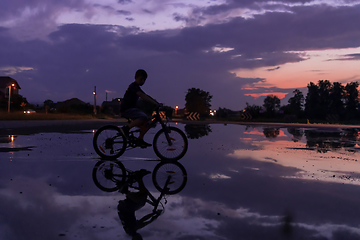 Image showing Lonely children silhouette on bike, boy riding bicycle on reflective water. Background beautiful sunset.