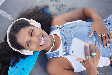 Image showing Portrait, phone and music with a black woman skater lying on her board at a skatepark from above. Face, skating and social media with a female skateboarder resting on the ground during a break