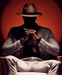 Image showing Stylish, fashionable and trendy man in silhouette sitting in a dark room with a red studio background. Edgy, mysterious and curious male mafia boss sitting in a fancy suit, hat and expensive jewelry