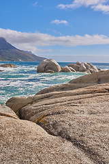 Image showing Beautiful sea view of a big boulders and ocean water on a sunny beach day in summer. A seaside setting in nature with a blue sky, white clouds, and waves. A seascape near a shore and mountains.