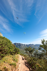 Image showing Nature park landscape with plants and an outdoor dirt path. Beautiful outdoors view of a hiking trail in the mountains during summer. Mountain background with a blue sky and paragliding travelers.