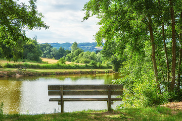 Image showing Relaxing nature view of a park bench with trees, grass, and a lake in the background. A forest and field landscape in the countryside. Natural outdoor setting perfect for a summer day outside