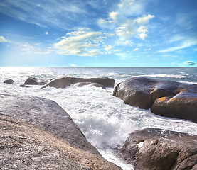 Image showing Copy space ocean view of beach with foamy sea water crashing onto boulders and rocks on a peaceful summer vacation in South African. Texture and detail of scenic coastline and cloudy blue sky