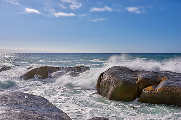 Image showing Beautiful landscape view of the rough ocean waves breaking on large boulders with a blue sky background. Tidal waves crashing into rocks on the windy wild coast and beach of Cape Town, South Africa