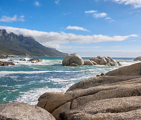 Image showing A calming ocean view of the rocky coast of Camps Bay in Cape Town, South Africa. Boulders at a beach with the tide and current coming in and Table Mountain on the horizon. Tidal waves washing ashore