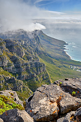 Image showing Clouds and fog covering a mountain peak on a sunny day along the coast. Scenic landscape with beautiful views of a rocky terrain in nature. Calm sea around an iconic landmark and travel destination