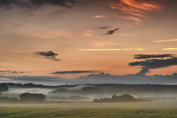 Image showing Beautiful sunrise with mist against a dramatic and colorful sky background. Calm and breathtaking landscape view on a field in the countryside with blooming trees on a foggy and overcast morning