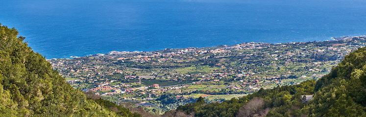 Image showing Landscape of a coastal city between hills and mountains from above. A peaceful village beside calm blue ocean water in the Canary Islands. High angle view of Los Llanos, La Palma in summer