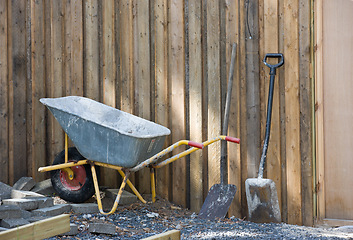 Image showing A wheelbarrow and spade placed amongst gravel and building material. Construction site at a home in the backyard. Equipment and tools used to build a house. A shovel and other building equipment