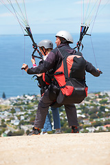 Image showing People, paragliding and outdoor to land on ground, extreme sport and sunshine by ocean on hill. Coach, person and back on adventure with helmet, backpack or parachute for jump, safety and blue sky
