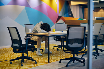 Image showing In a modern, spacious startup office, an exhausted African American man lies on a desk, reflecting the challenges of contemporary corporate life and the importance of work-life balance.