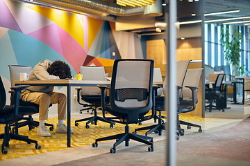 Image showing In a modern, spacious startup office, an exhausted African American man lies on a desk, reflecting the challenges of contemporary corporate life and the importance of work-life balance.