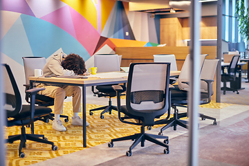 Image showing In a modern, spacious startup office, an exhausted African American man lies on a desk, reflecting the challenges of contemporary corporate life and the importance of work-life balance.